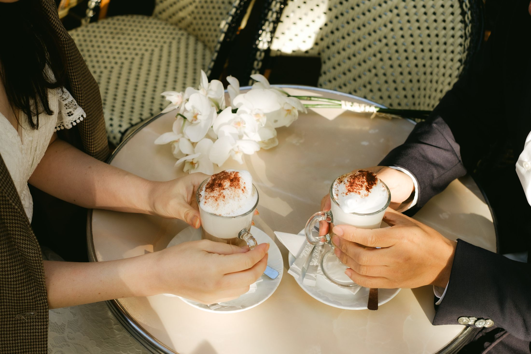 Zwei Hände halten Cappuccino-Gläser an einem Marmortisch mit Orchideen